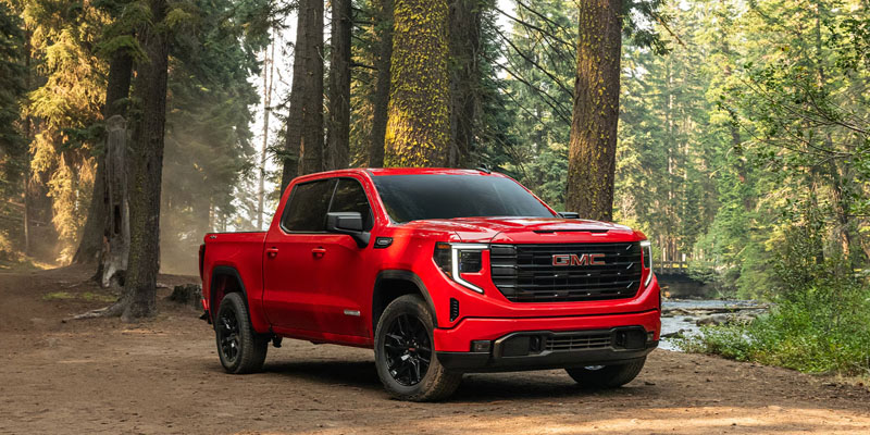 A red 2026 GMC Sierra 1500 parked on a dirt space under the forest park trees beside a rocky creek.