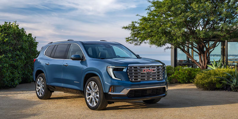 A soft blue-colored 2026 GMC Acadia SUV parked beside green tree and shrubs at a lake.