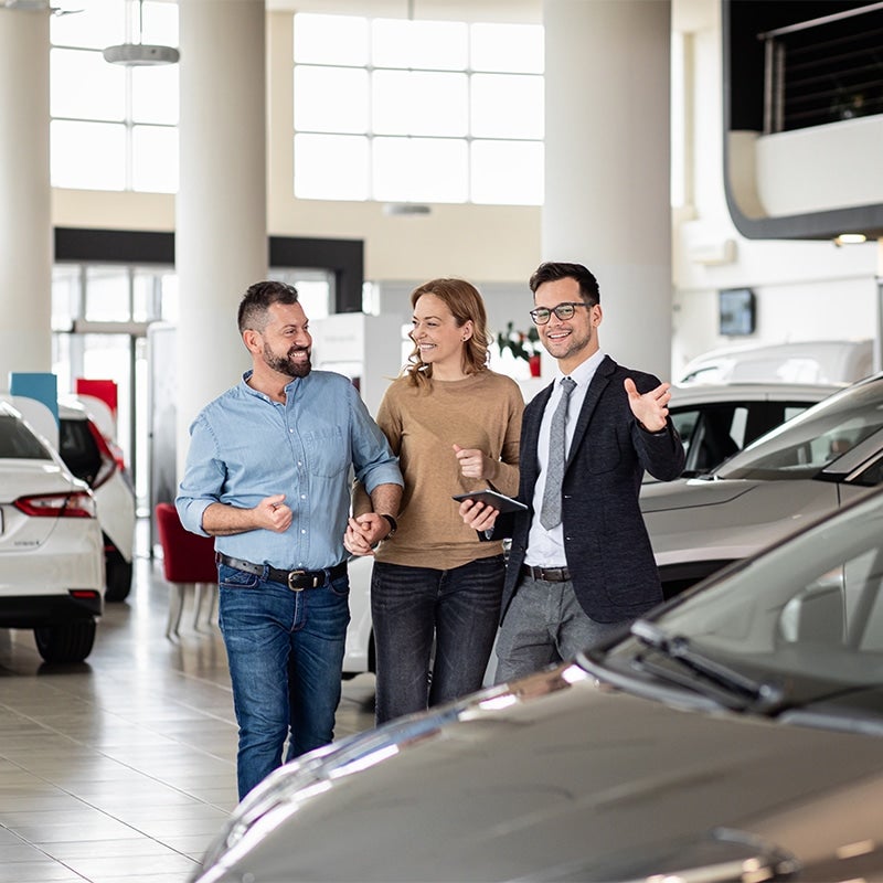 A smiling, dark-suited GMC salesperson shows an excited couple who are holding hands and smiling at one another arouind the bright, white showroom.