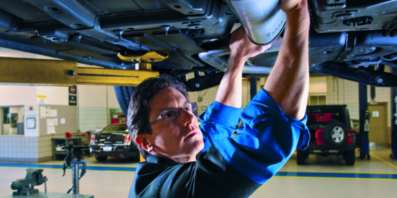 A blue-suited certified GMC Service Center technician is inspecting and repairing a GMC vehicle raised above him.